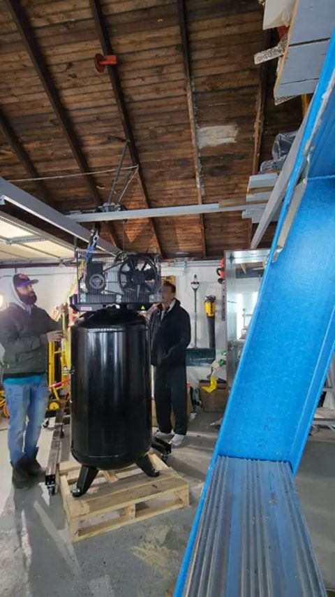 two men moving a large upright air compressor on a pallet in a garage, face visible