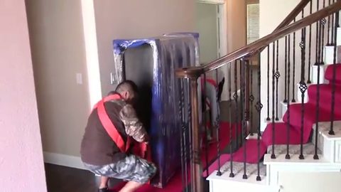 two movers hoisting a shrink-wrapped dresser up interior stairs using lifting straps