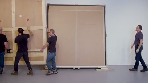 art handlers opening a wooden shipping crate for a large framed painting in a gallery, face visible