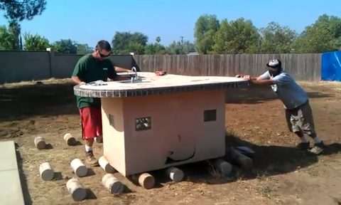crew moving a large outdoor kitchen BBQ island on pipe rollers, face visible