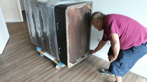 mover maneuvering a plastic-wrapped dresser on a dolly into an apartment, face visible